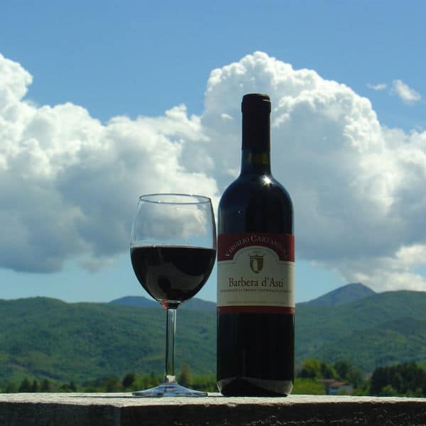 Una bottiglia di Barbera d'Asti DOCG ed un bicchiere di vino rosso si stagliano in controluce su uno sfondo con verdi colline e un cielo azzurro con cumuli di nubi bianche
