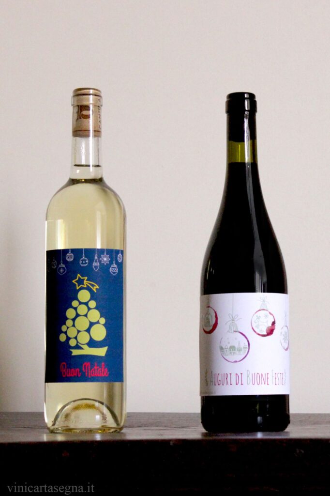 Bottiglie di vino con etichette personalizzate per la tavola delle feste: l'etichetta sulla bottiglia di vino bianco ha lo sfondo blu con un grappolo d'uva verde capovolto, che sembra un albero di natale, e la scritta in rosso "Buon Natale". L'etichetta della bottiglia del vino rosso è bianca con 4 cerchi che sono l'impronta del fondo di un bicchiere di vino rosso e che sembrano palline di natale. Sotto, c'è la scritta "Auguri di Buone Feste"